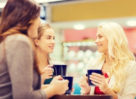 drinks, communication, friendship and people concept - happy young women with cups sitting at table and talking in mall or cafe